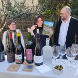 Trois personnes souriantes devant une table garnie de bouteilles de vin, de verres et d'un magazine.