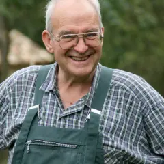 Henri Bonneau, un homme souriant avec des lunettes, porte une salopette verte et une chemise à carreaux.