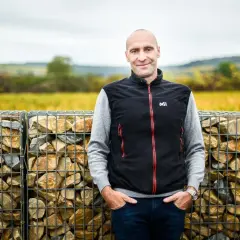 Pierre Yves Colin Morey, vigneron souriant, pose devant des murs de gabions remplis de pierres, avec des vignes en arrière-plan.