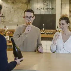 Dégustation de bulles chez Ackerman à Saumur. Un homme et une femme savourent un verre, tandis qu'un serveur présente une bouteille.