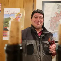 Un vigneron du domaine des Nugues présente un verre de vin rouge, avec des cartes du Beaujolais en arrière-plan.