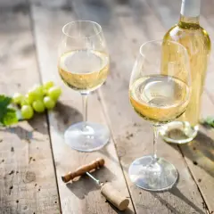 Deux verres de vin blanc sec, une bouteille, des raisins verts et un tire-bouchon sur une table en bois.