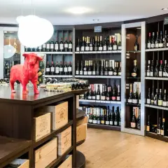 Une cave à vin bien garnie, avec des étagères remplies de bouteilles. Au premier plan, deux grandes bouteilles de champagne. Un taureau rouge trône sur le comptoir, clin d'œil aux 20 meilleurs vins du Languedoc.