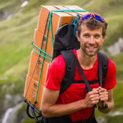 François d'Haene, vigneron, porte une lourde charge de caisses de vin sur son dos, traversant un paysage montagneux verdoyant.