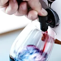 Un doigt appuie sur le robinet d'un cubi de vin blanc ouvert, versant le liquide rouge dans un verre.