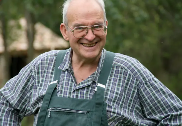 Henri Bonneau, un homme souriant avec des lunettes, porte une salopette verte et une chemise à carreaux.
