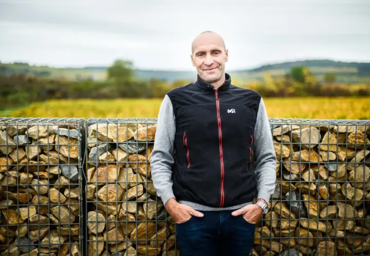Pierre Yves Colin Morey, vigneron souriant, pose devant des murs de gabions remplis de pierres, avec des vignes en arrière-plan.