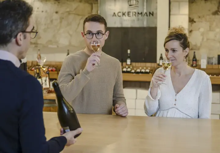 Dégustation de bulles chez Ackerman à Saumur. Un homme et une femme savourent un verre, tandis qu'un serveur présente une bouteille.