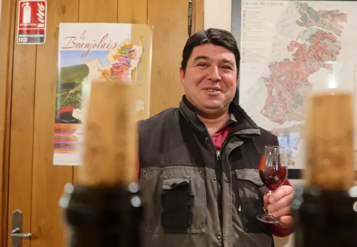 Un vigneron du domaine des Nugues présente un verre de vin rouge, avec des cartes du Beaujolais en arrière-plan.