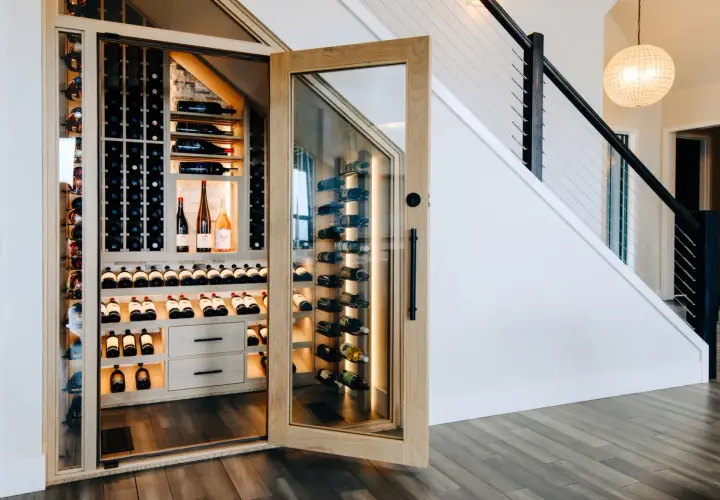 Belle cave à vin moderne sous un escalier, avec des étagères en bois et des bouteilles illuminées.