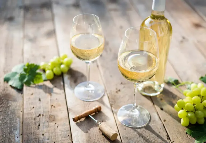 Deux verres de vin blanc sec, une bouteille, des raisins verts et un tire-bouchon sur une table en bois.