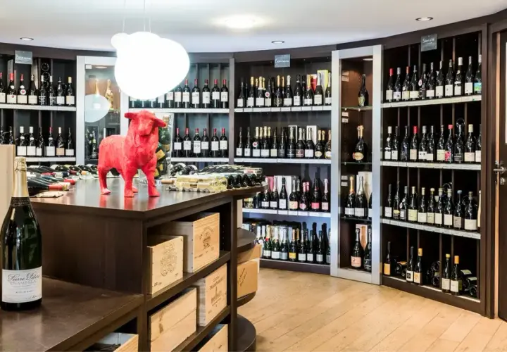 Une cave à vin bien garnie, avec des étagères remplies de bouteilles. Au premier plan, deux grandes bouteilles de champagne. Un taureau rouge trône sur le comptoir, clin d'œil aux 20 meilleurs vins du Languedoc.