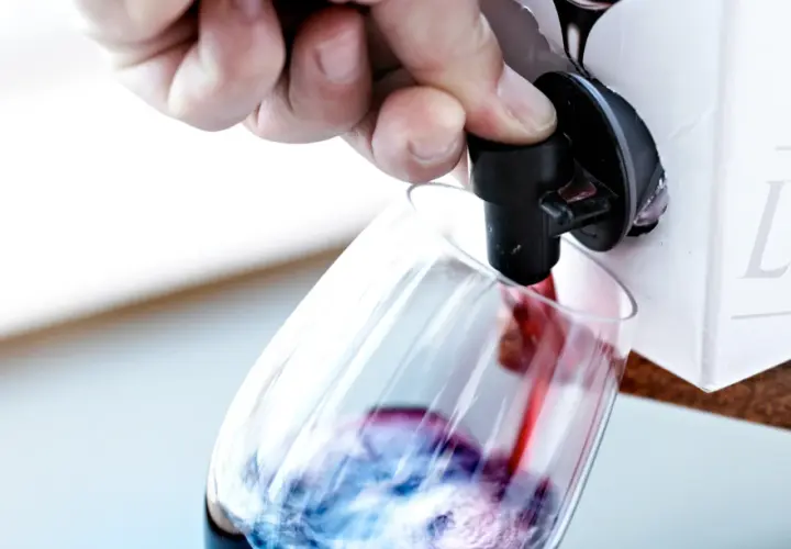 Un doigt appuie sur le robinet d'un cubi de vin blanc ouvert, versant le liquide rouge dans un verre.