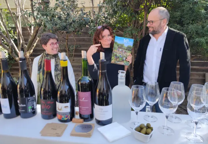 Trois personnes souriantes devant une table garnie de bouteilles de vin, de verres et d'un magazine.