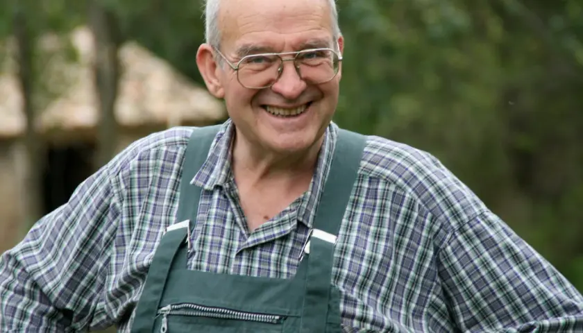 Henri Bonneau, un homme souriant avec des lunettes, porte une salopette verte et une chemise à carreaux.
