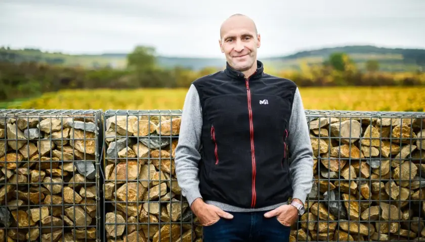 Pierre Yves Colin Morey, vigneron souriant, pose devant des murs de gabions remplis de pierres, avec des vignes en arrière-plan.