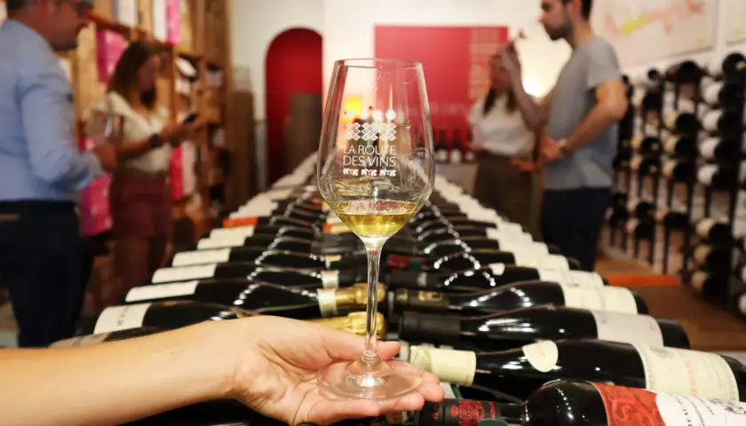 Verre de vin blanc sur fond de bouteilles alignées, évoquant les plaisirs de la route des vins.