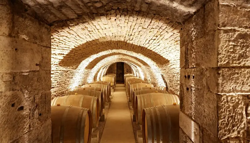 Cave voûtée en pierre, éclairée par des spots, remplie de fûts de chêne. Un lieu idéal pour le vieillissement des vins de Prosper Maufoux.