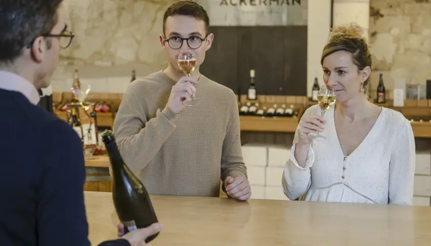 Dégustation de bulles chez Ackerman à Saumur. Un homme et une femme savourent un verre, tandis qu'un serveur présente une bouteille.