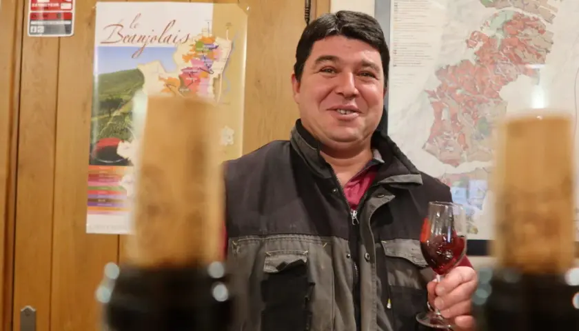 Un vigneron du domaine des Nugues présente un verre de vin rouge, avec des cartes du Beaujolais en arrière-plan.