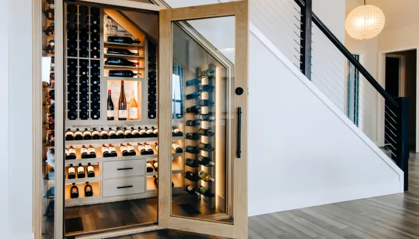 Belle cave à vin moderne sous un escalier, avec des étagères en bois et des bouteilles illuminées.