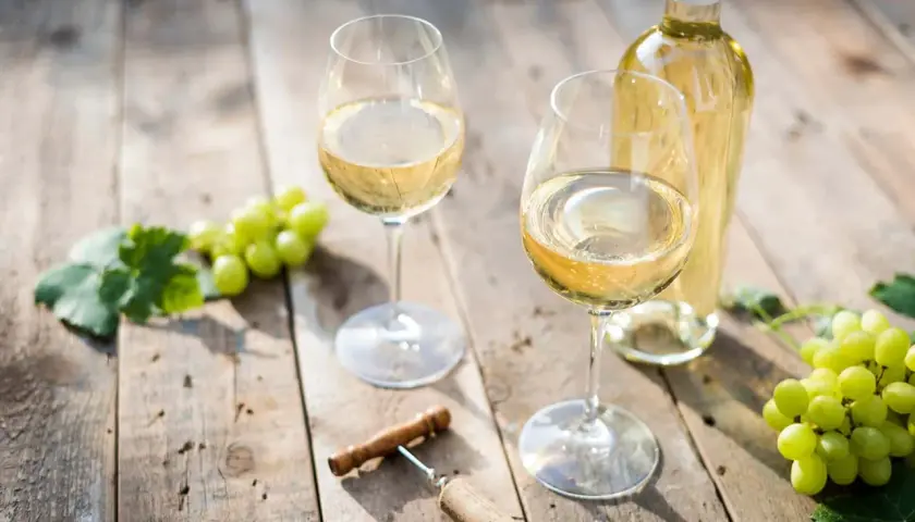 Deux verres de vin blanc sec, une bouteille, des raisins verts et un tire-bouchon sur une table en bois.