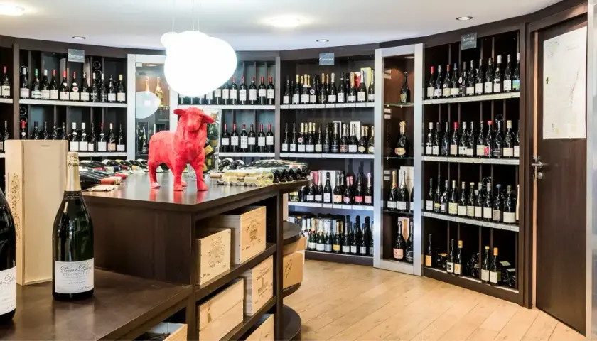 Une cave à vin bien garnie, avec des étagères remplies de bouteilles. Au premier plan, deux grandes bouteilles de champagne. Un taureau rouge trône sur le comptoir, clin d'œil aux 20 meilleurs vins du Languedoc.