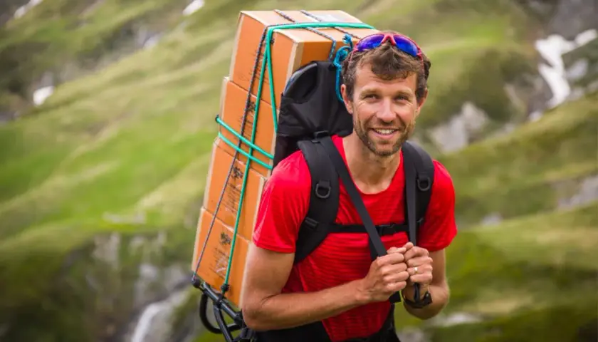 François d'Haene, vigneron, porte une lourde charge de caisses de vin sur son dos, traversant un paysage montagneux verdoyant.