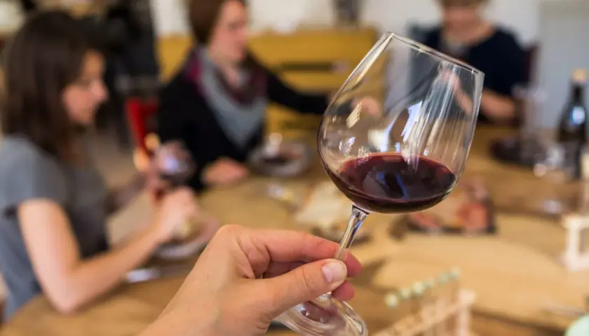 Une main tient un verre de vin rouge, prêt pour une initiation oenologie. Des personnes sont assises à une table en arrière-plan.
