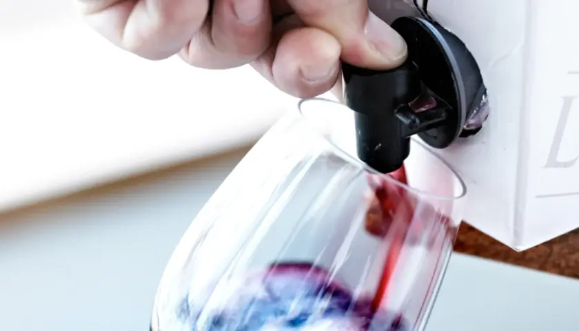 Un doigt appuie sur le robinet d'un cubi de vin blanc ouvert, versant le liquide rouge dans un verre.