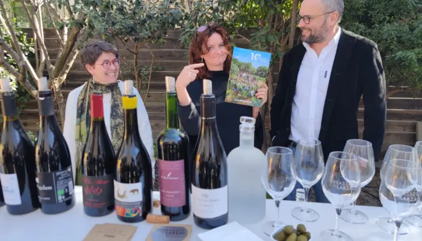 Trois personnes souriantes devant une table garnie de bouteilles de vin, de verres et d'un magazine.