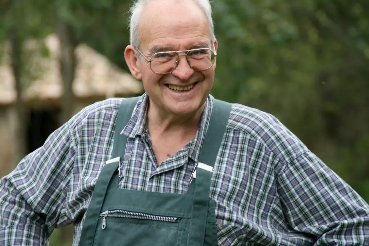 Henri Bonneau, un homme souriant avec des lunettes, porte une salopette verte et une chemise à carreaux.
