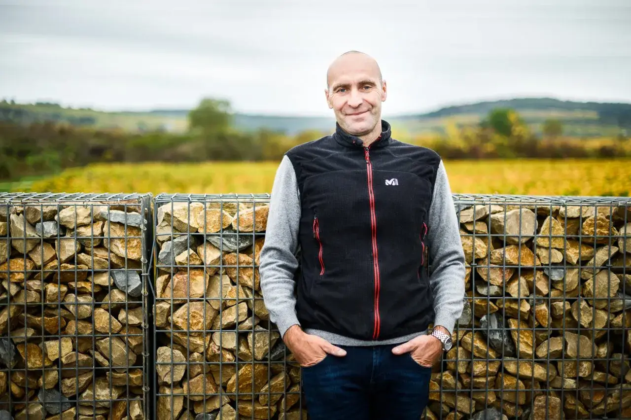 Pierre Yves Colin Morey, vigneron souriant, pose devant des murs de gabions remplis de pierres, avec des vignes en arrière-plan.