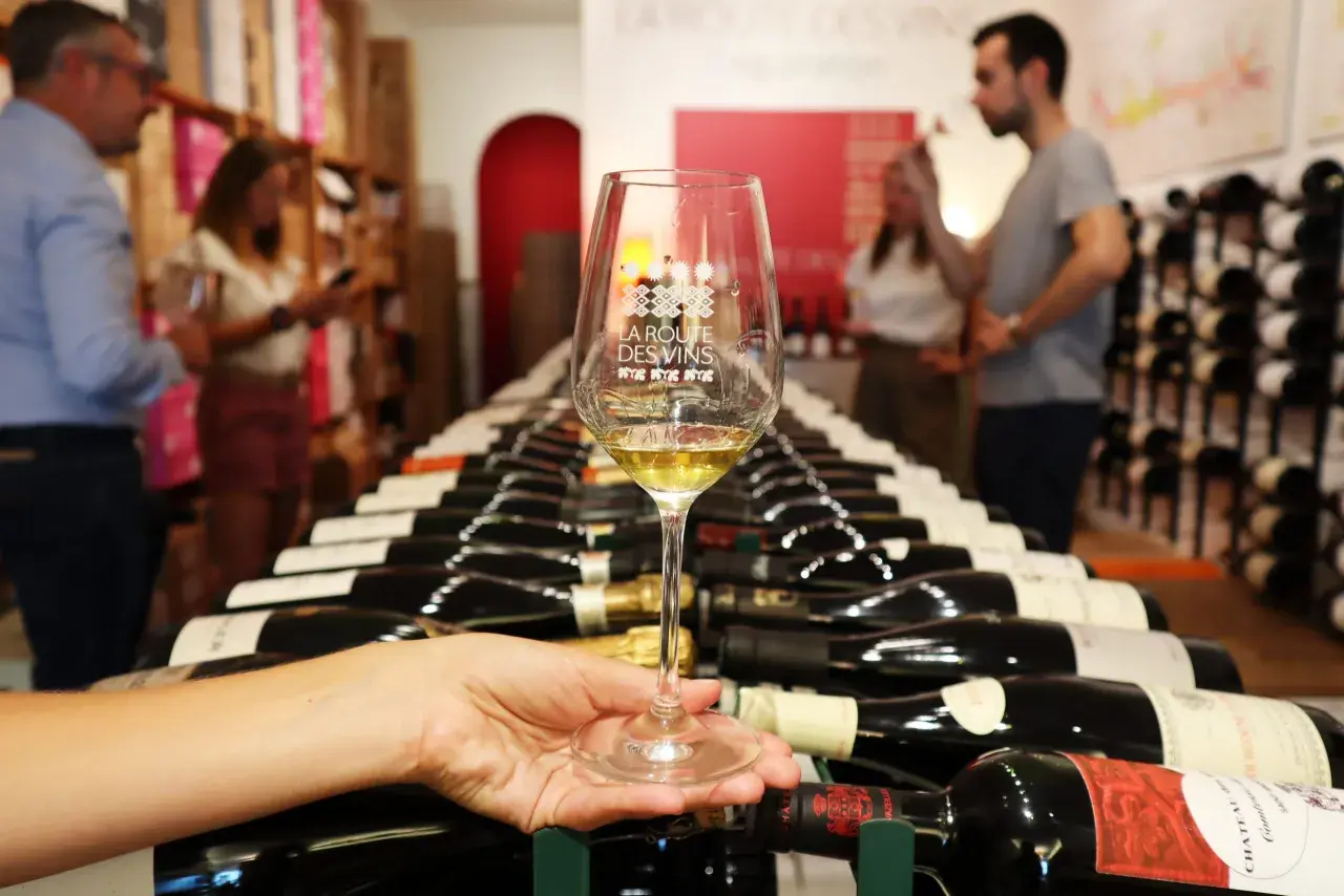 Verre de vin blanc sur fond de bouteilles alignées, évoquant les plaisirs de la route des vins.