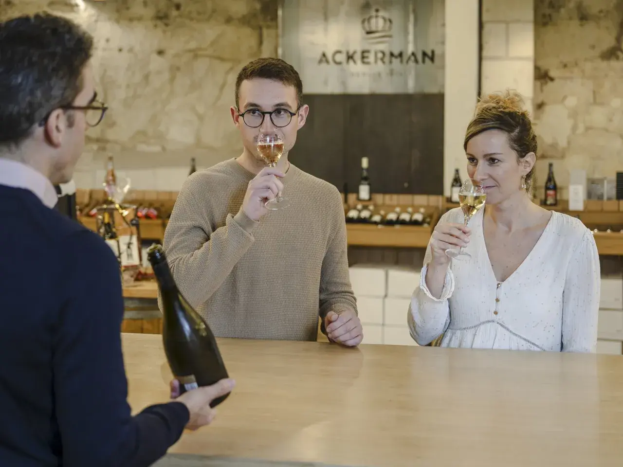 Dégustation de bulles chez Ackerman à Saumur. Un homme et une femme savourent un verre, tandis qu'un serveur présente une bouteille.