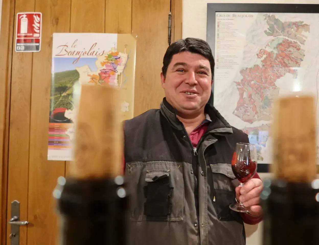 Un vigneron du domaine des Nugues présente un verre de vin rouge, avec des cartes du Beaujolais en arrière-plan.