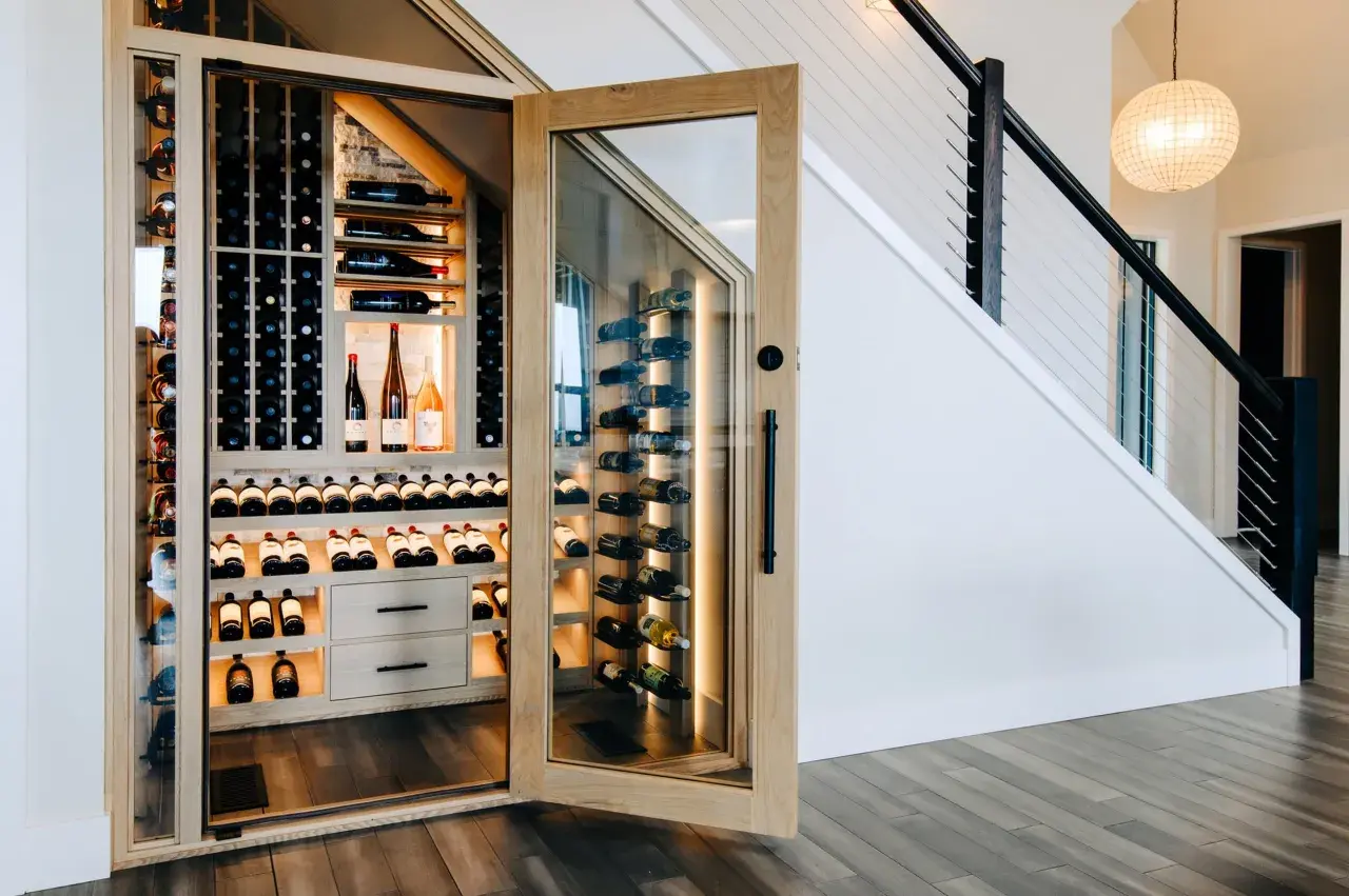 Belle cave à vin moderne sous un escalier, avec des étagères en bois et des bouteilles illuminées.