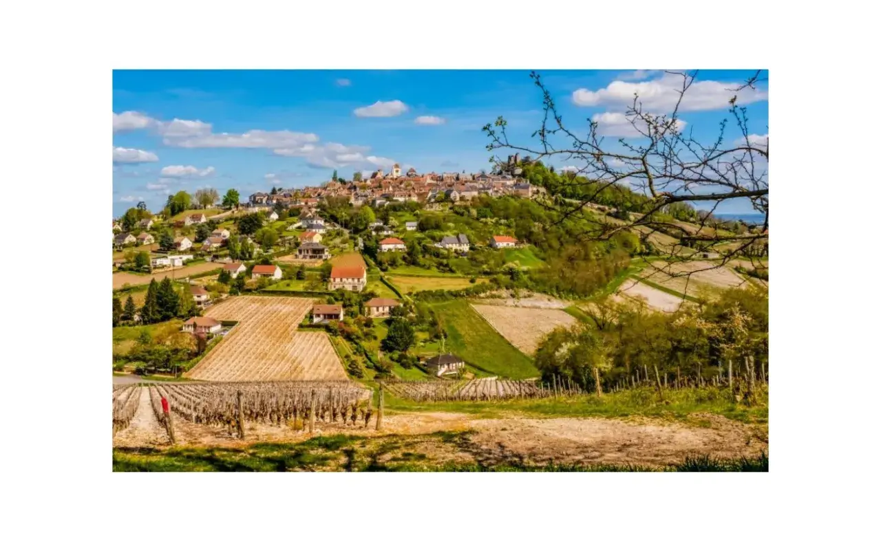 Paysage bucolique de la région de Sancerre, avec un village perché sur une colline, entouré de vignobles et de champs verdoyants sous un ciel bleu.