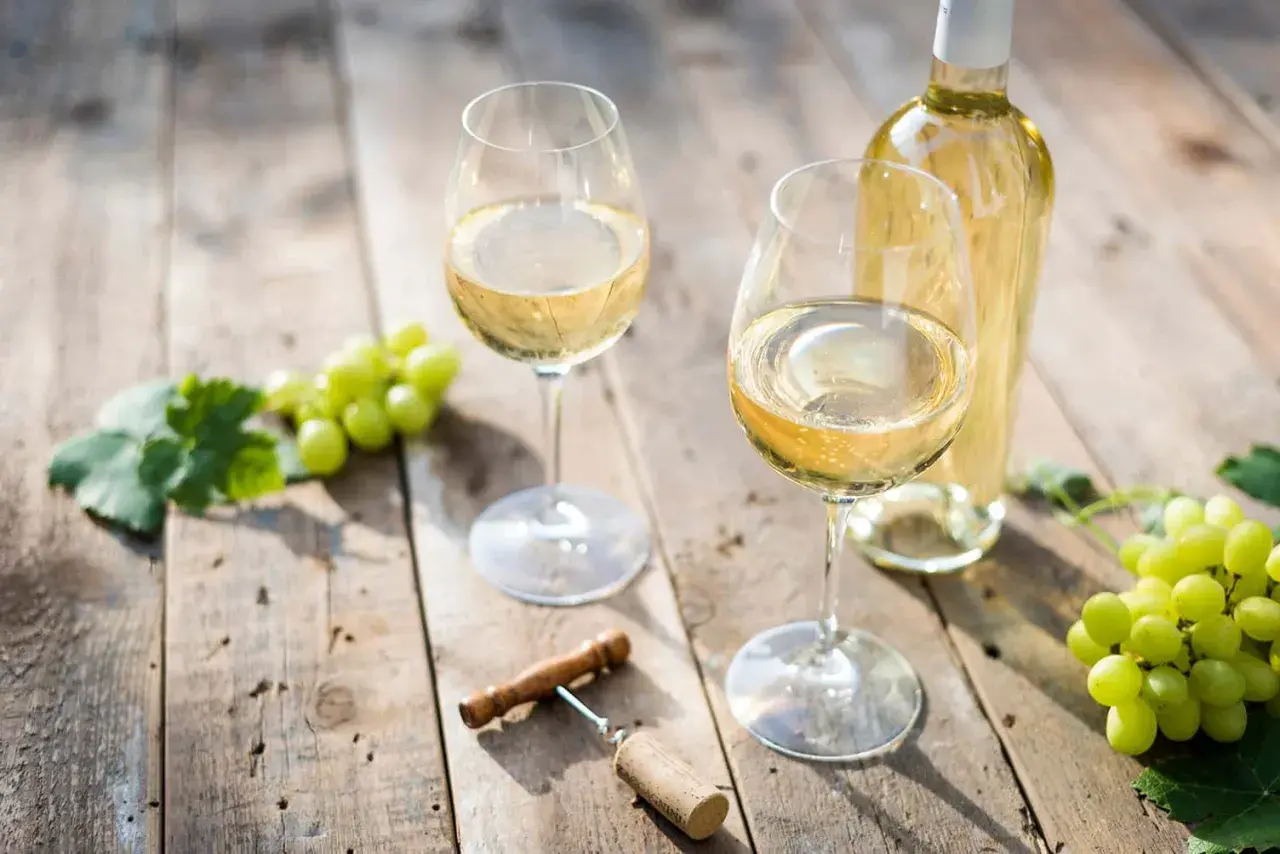 Deux verres de vin blanc sec, une bouteille, des raisins verts et un tire-bouchon sur une table en bois.