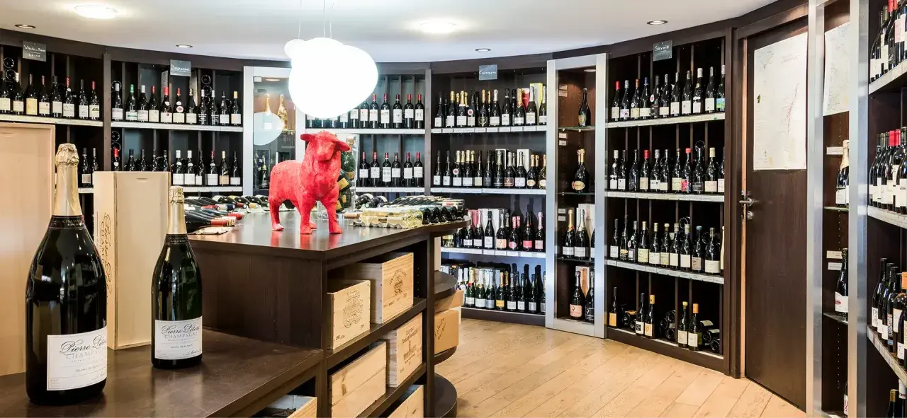 Une cave à vin bien garnie, avec des étagères remplies de bouteilles. Au premier plan, deux grandes bouteilles de champagne. Un taureau rouge trône sur le comptoir, clin d'œil aux 20 meilleurs vins du Languedoc.