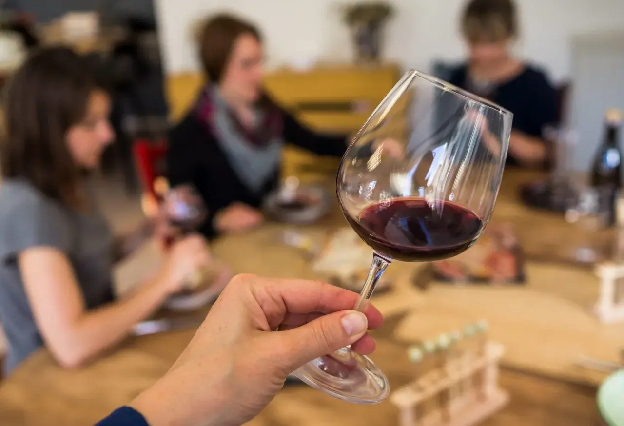 Une main tient un verre de vin rouge, prêt pour une initiation oenologie. Des personnes sont assises à une table en arrière-plan.