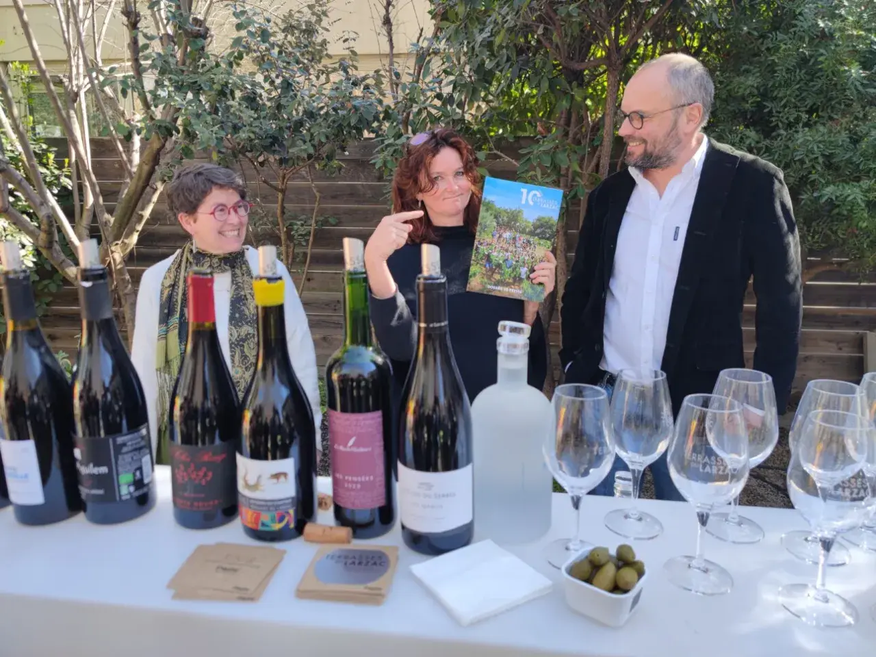 Trois personnes souriantes devant une table garnie de bouteilles de vin, de verres et d'un magazine.