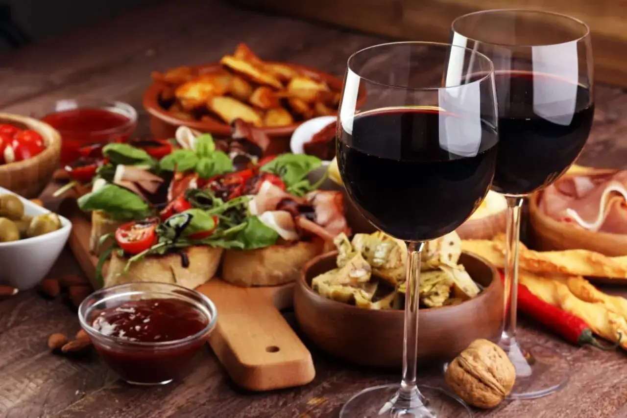 Deux verres de vin rouge accompagnent un assortiment de tapas : bruschettas, artichauts, olives, frites et charcuterie. Une invitation à un moment de