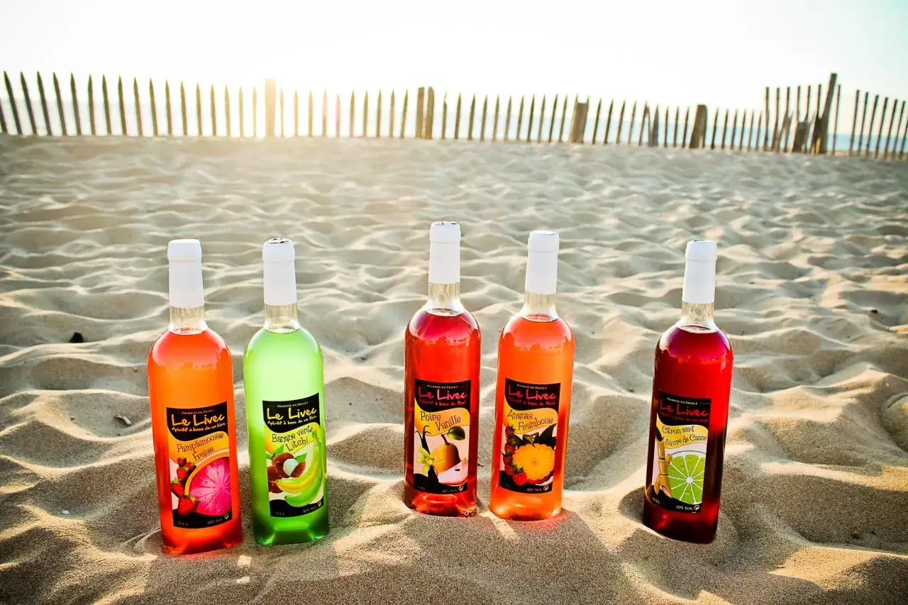 Cinq bouteilles d'ap&eacute;ritif &agrave; base de vin blanc aux saveurs fruit&eacute;es, pos&eacute;es sur le sable, parfaites pour un ap&eacute;ro au coucher du soleil.
