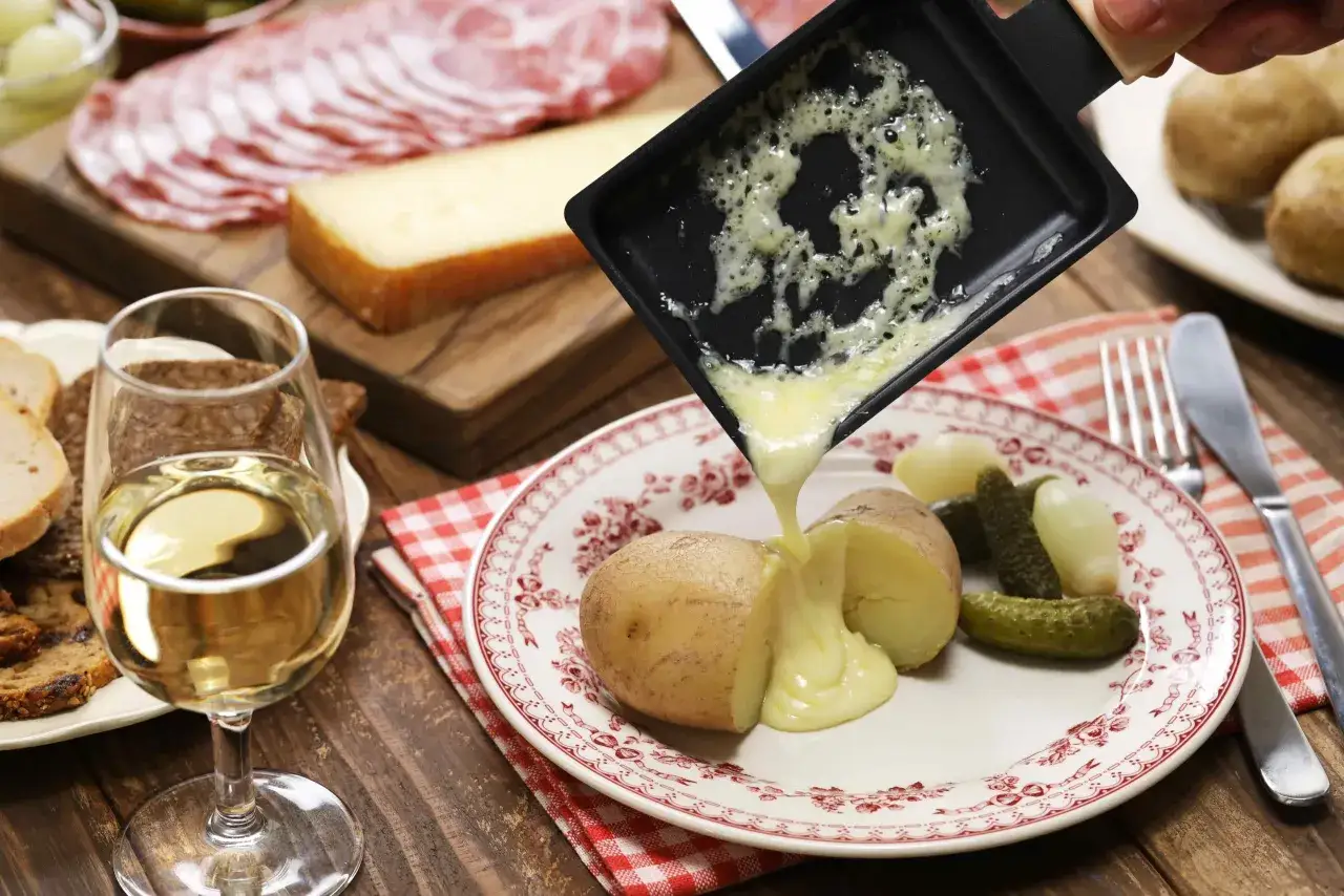 Raclette fondue versée sur des pommes de terre, accompagnée de cornichons et d'un verre de vin blanc, parfait avec un Pouilly Fumé.
