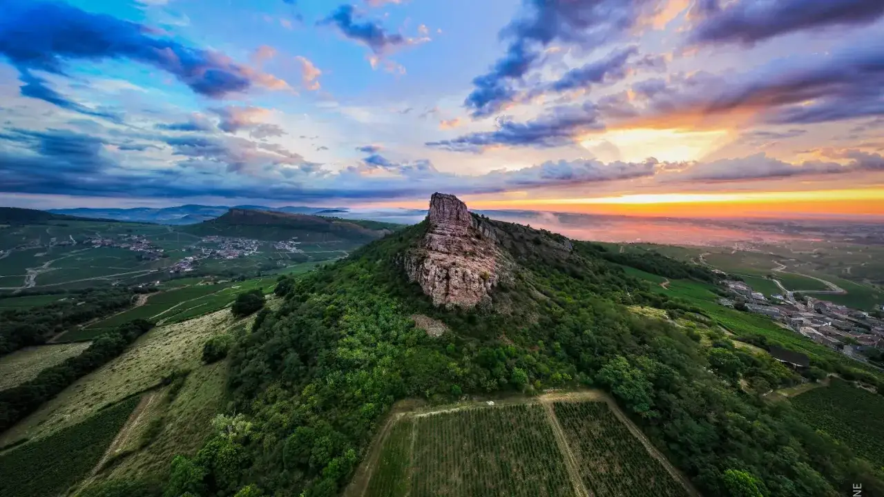 Le rocher de Solutr&eacute;, un site embl&eacute;matique de la Bourgogne, se dresse fi&egrave;rement au milieu des vignobles verdoyants. Le soleil couchant, avec ses teintes orang&eacute;es et roses, illumine le ciel, cr&eacute;ant une atmosph&egrave;re magique. Ce paysage, immortalis&eacute; par Guf...