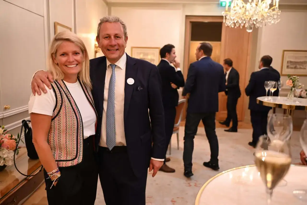 Alain Marty pose avec une femme souriante lors d'une réception élégante, sous un lustre scintillant.