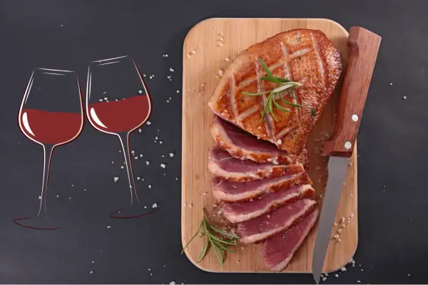 Un plat de magret de canard tranché, garni de romarin, prêt à être dégusté avec deux verres de vin rouge.